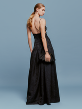 Load image into Gallery viewer, A woman stands against a plain blue background, wearing the Maxine Dress Black—a long dress with thin straps and beautiful floral detailing. She holds a small black purse and wears a long necklace, capturing an elegant style reminiscent of late summer evenings between August 19th and September 2nd, 2024.
