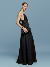 Load image into Gallery viewer, A woman stands against a plain blue background, wearing the Maxine Dress Black—a long dress with thin straps and beautiful floral detailing. She holds a small black purse and wears a long necklace, capturing an elegant style reminiscent of late summer evenings between August 19th and September 2nd, 2024.
