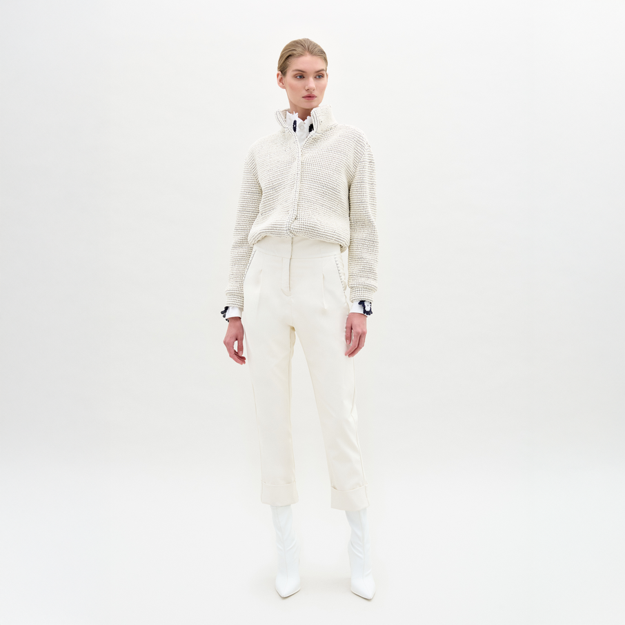 A person stands against a plain light background, wearing a textured white sweater, white collared shirt, the Moad Pant—an Italian fabric high-waisted cropped pant—and white heeled boots.