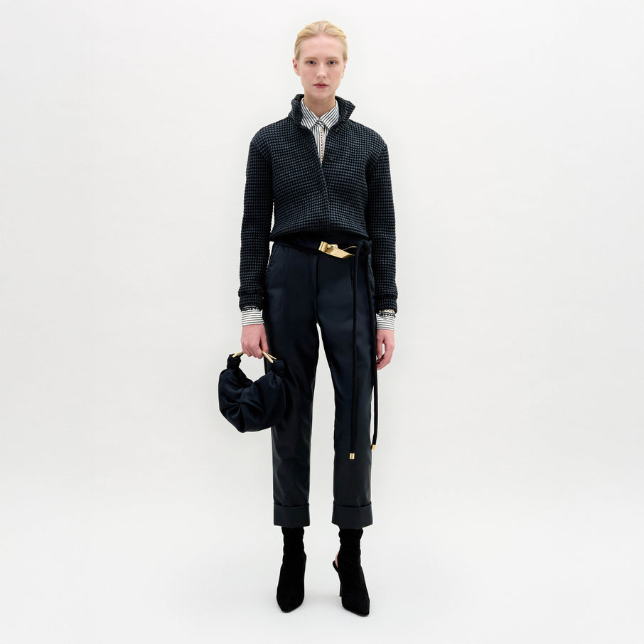 Person stands against a plain white background wearing the Moad Pant, a dark textured jacket, patterned shirt, black boots, and holding a small black handbag.