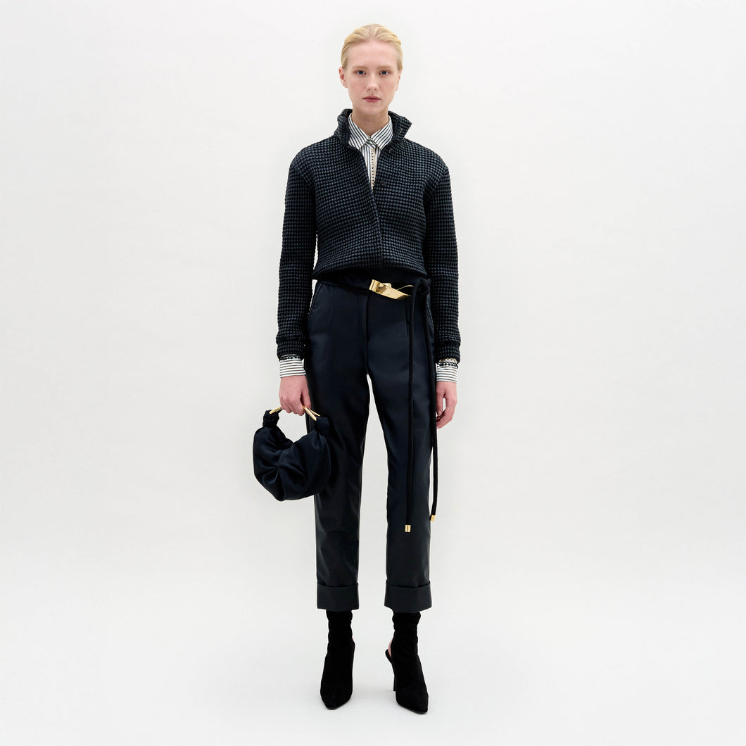 Person stands against a plain white background wearing the Moad Pant, a dark textured jacket, patterned shirt, black boots, and holding a small black handbag.