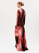 Load image into Gallery viewer, A woman with blonde hair in a neat bun faces away, modeling the Maialen Dress—a long-sleeved, floor-length gown featuring a pink and brown tie-dye pattern—set against a plain white background.
