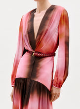 Load image into Gallery viewer, A person wears the Maialen Dress—a long-sleeved, deep V-neck, pink and brown gradient dress with a gathered waist and braided red belt with gold accents. The photo is cropped at the shoulders and mid-thigh.
