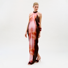 Load image into Gallery viewer, A woman stands against a plain light background wearing the Mene Dress, a flowing halter-neck gown in pink, orange, and brown tones. Her hair is pulled back, and she accessorizes with a maroon bracelet and matching heeled sandals.
