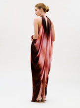 Load image into Gallery viewer, A woman with blonde hair in a low bun wears the Mene Dress—a flowing, sleeveless pink and brown halter-neck gown—standing sideways and glancing over her shoulder against a plain light background.

