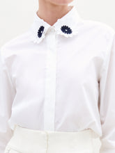 Load image into Gallery viewer, A woman wears the Naroa Blouse—a white top with floral appliqué details—paired with a white Italian-fabric skirt, standing against a plain light background.
