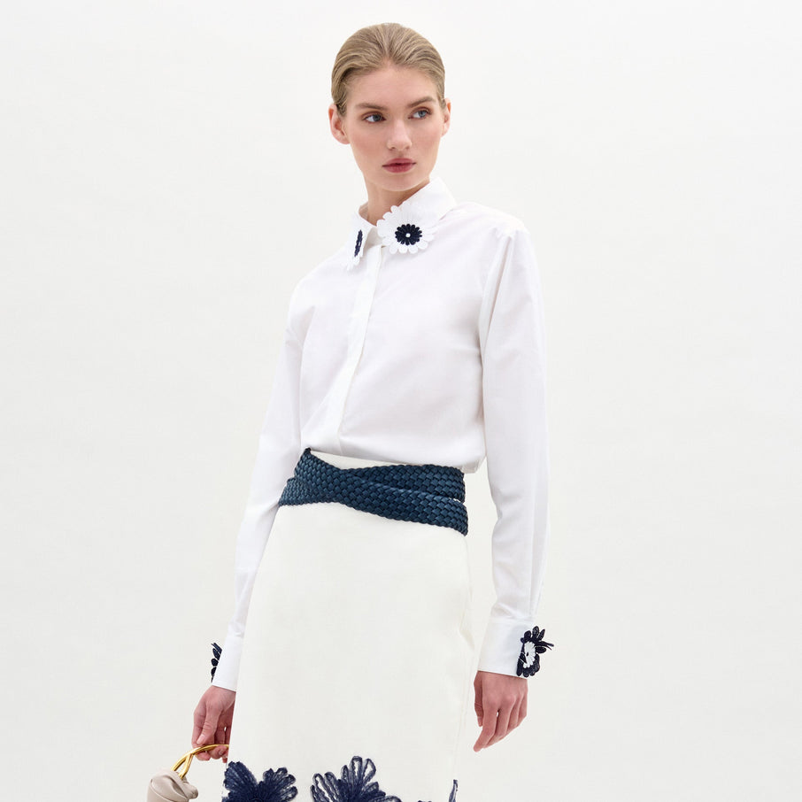 A woman wears the Naroa Blouse—a white top with floral appliqué details—paired with a white Italian-fabric skirt, standing against a plain light background.