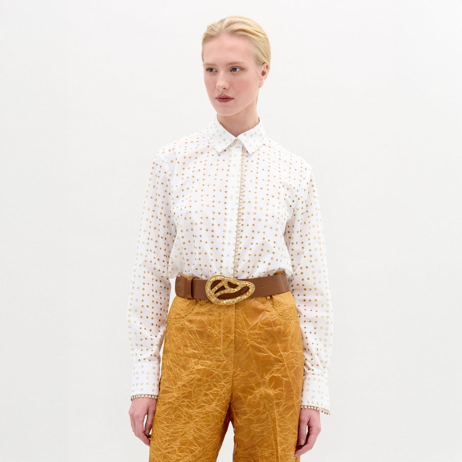 Person wearing a white patterned shirt and mustard yellow pants with a brown belt on a light gray background