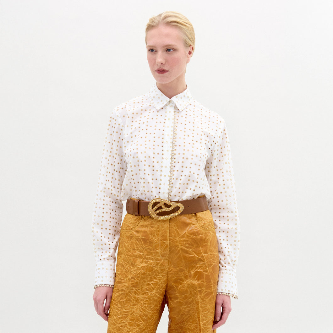 Person wearing a white patterned shirt and mustard yellow pants with a brown belt on a light gray background