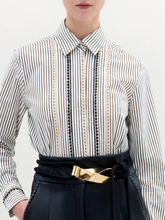 Load image into Gallery viewer, Person stands against a plain background, wearing the Naroa Blouse— a striped 100% cotton button-up— tucked into high-waisted black pants with a gold-buckle belt.
