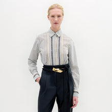 Load image into Gallery viewer, Person stands against a plain background, wearing the Naroa Blouse— a striped 100% cotton button-up— tucked into high-waisted black pants with a gold-buckle belt.
