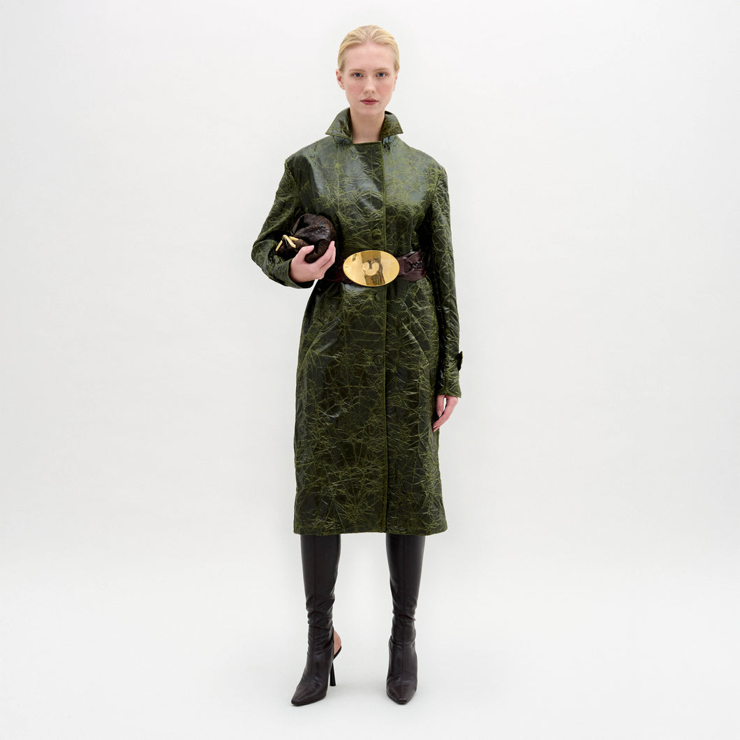 A woman wears the Nilo Trench, a textured dark green coat crafted from Italian fabric, paired with dark boots, a large gold belt buckle, and holding a small dark clutch against a plain background.