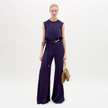 Load image into Gallery viewer, A woman wearing the Noel Jumpsuit in color Purple with a Turni clutch in color Camel on a white background.
