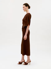 Load image into Gallery viewer, A woman stands in profile against a plain light background, wearing the Oby Dress—a brown short-sleeved midi dress crafted from luxurious Italian fabric—paired with brown heeled sandals. Her hair is tied back in a neat bun.
