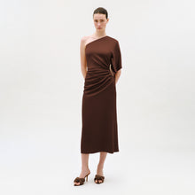 Load image into Gallery viewer, A woman stands against a white background, wearing the Oby Dress—an elegant midi dress with an asymmetric neckline, crafted from luxurious Italian fabric. The brown, one-shoulder design features side ruching and is paired with matching brown heeled sandals. Her hair is pulled back and her hands are behind her back.
