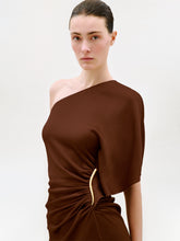 Load image into Gallery viewer, A woman with dark hair in a sleek bun wears the Oby Dress, a brown one-shoulder midi dress crafted from luxurious Italian fabric with a gold accent at the waist, standing against a plain light background.
