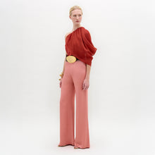 Load image into Gallery viewer, A woman stands in high-waisted pink Palermo Pants, crafted from luxurious Italian fabric, paired with a red one-shoulder top, gold belt, gold bracelets, and open-toe shoes against a white background.
