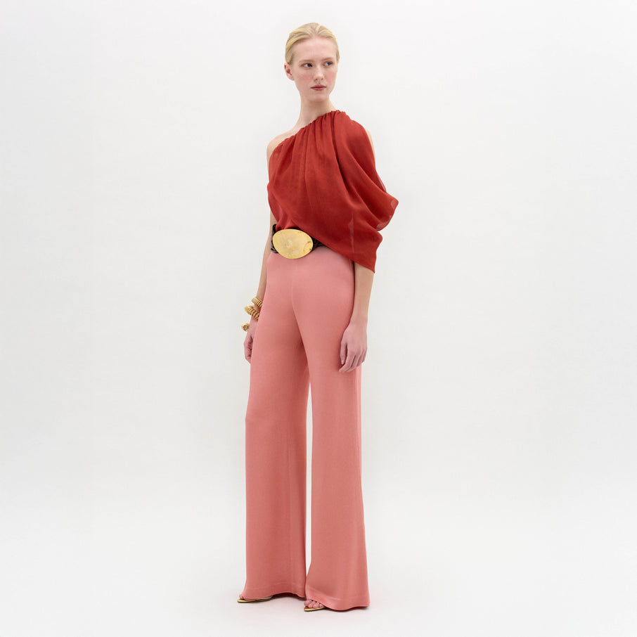 A woman stands in high-waisted pink Palermo Pants, crafted from luxurious Italian fabric, paired with a red one-shoulder top, gold belt, gold bracelets, and open-toe shoes against a white background.