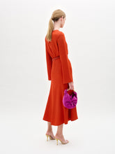 Load image into Gallery viewer, Woman wearing the Pesaro Dress, a long-sleeved orange midi dress with a belted waist crafted from premium Italian fabric, paired with a small purple bag and transparent high-heeled sandals, against a plain white background.
