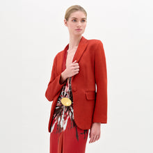 Load image into Gallery viewer, A person wearing the Rebeca Jacket, a fitted red blazer made from Italian fabric, layered over a patterned red and beige dress, stands against a plain light background.
