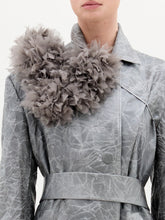 Load image into Gallery viewer, A woman stands against a plain background wearing the Nilo Trench, a textured gray structured coat with a large floral shoulder detail, paired with black heeled boots and a gold ankle accessory.
