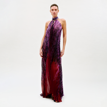 Load image into Gallery viewer, A person stands against a light background, wearing the Lucia Dress—a floor-length halter-neck gown featuring a purple and red abstract pattern with a metallic ring detail at the neckline.

