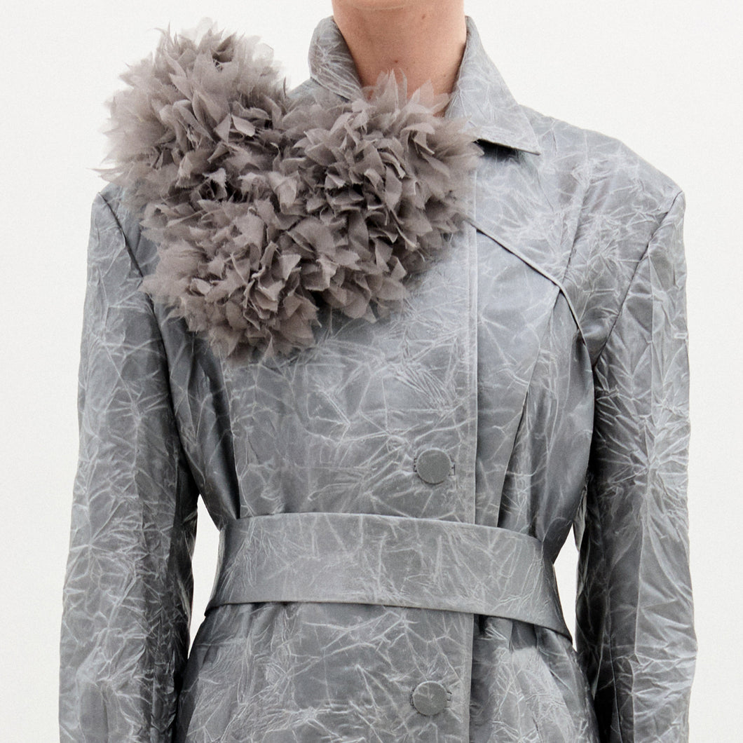 A person wears a light grey, textured coat made from Italian fabric, styled with the Romy Collar Piece—a large, ruffled, fluffy grey accessory attached near the left shoulder.