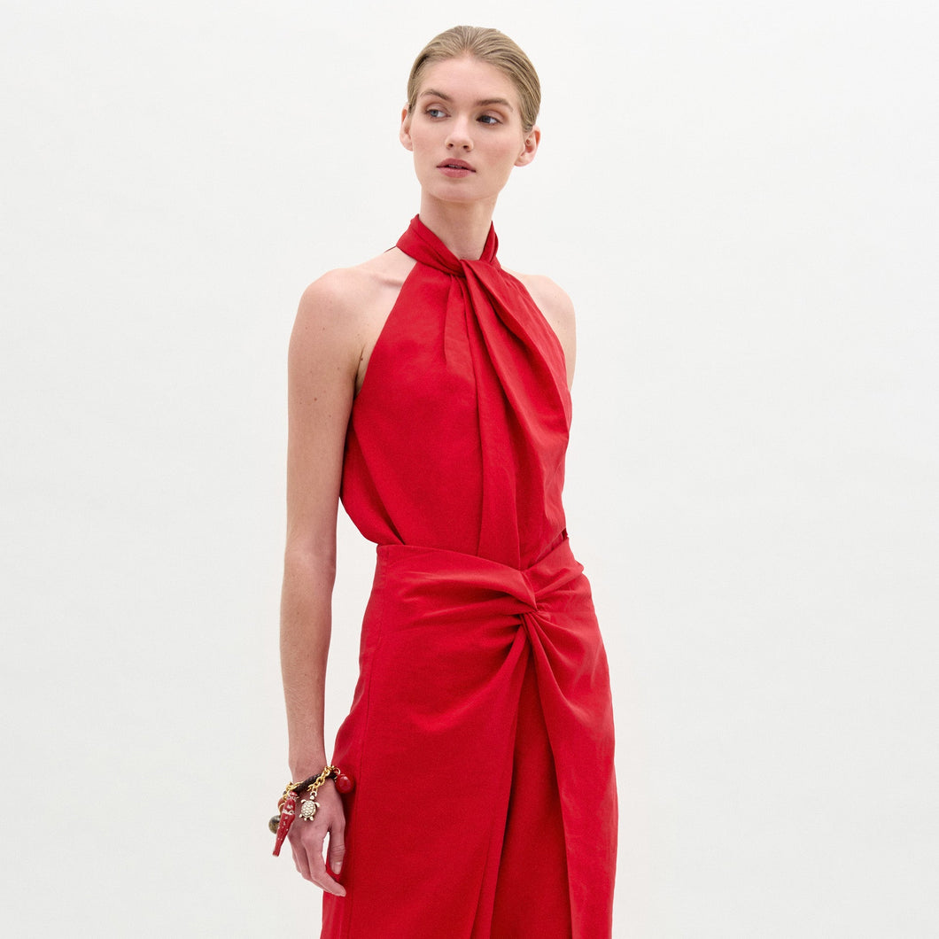 Woman wearing the Shela Blouse, a sleeveless red halter-neck top made from Italian fabric with a knotted front detail, standing against a plain light background.