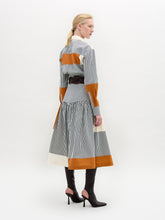 Load image into Gallery viewer, A woman models the Shoreh Skirt, featuring vertical stripes and orange color blocks in Italian fabric. She styles it with a wide gold-buckle belt and knee-high black boots, standing against a plain white background.

