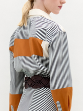 Load image into Gallery viewer, A person wearing the Soleil Blouse—long-sleeved and striped with orange and white color blocks in 100% Italian cotton—paired with a large gold belt, stands against a plain light background.
