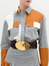 Load image into Gallery viewer, A person wearing the Soleil Blouse—long-sleeved and striped with orange and white color blocks in 100% Italian cotton—paired with a large gold belt, stands against a plain light background.
