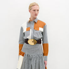 Load image into Gallery viewer, A person wearing the Soleil Blouse—long-sleeved and striped with orange and white color blocks in 100% Italian cotton—paired with a large gold belt, stands against a plain light background.
