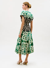 Load image into Gallery viewer, A woman stands slightly turned, wearing the Sakura Dress—a green and white floral midi dress with a black geometric waist detail—and beige heeled sandals. Her hair is styled in a neat bun.
