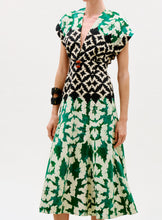 Load image into Gallery viewer, A woman wearing the Sakura Dress, featuring a geometric black and white bodice and green leaf print skirt, accessorized with chunky black bracelets, stands against a plain background.
