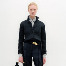 Load image into Gallery viewer, A person stands against a plain white background wearing the Zalta Jacket, a dark cropped piece crafted from Italian fabric, paired with a striped shirt, dark trousers with a gold belt, and holding a dark bag.
