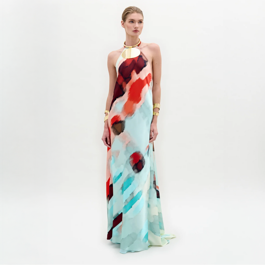 A woman poses against a white background wearing the Zeffa Dress, a sleeveless, floor-length silk maxi with an abstract red, aqua, and white pattern, paired with gold jewelry.